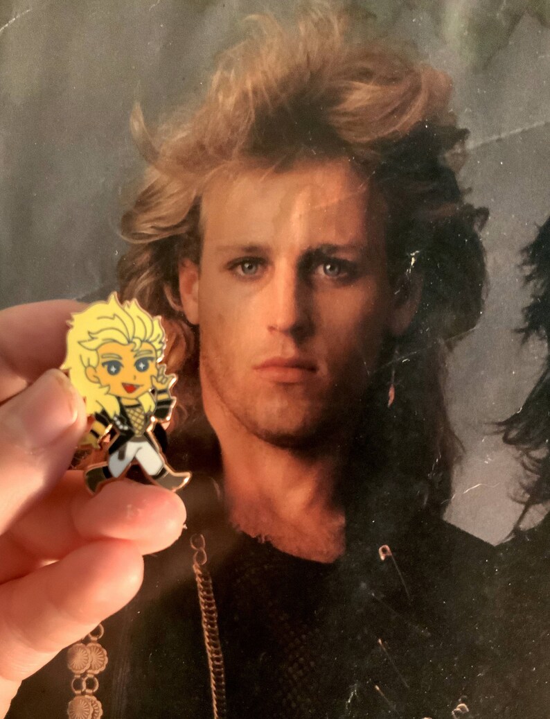 May include: A cartoon pin featuring a blonde character in a black and white outfit with gold accents. The pin is held in front of a vintage photograph of a man with blonde hair.