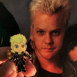 May include: A cartoon-style enamel pin featuring a character with blonde hair and a black outfit. The pin is held in front of a faded image of a man with blonde hair. The pin has a black outline and is filled with yellow, black, and red enamel.