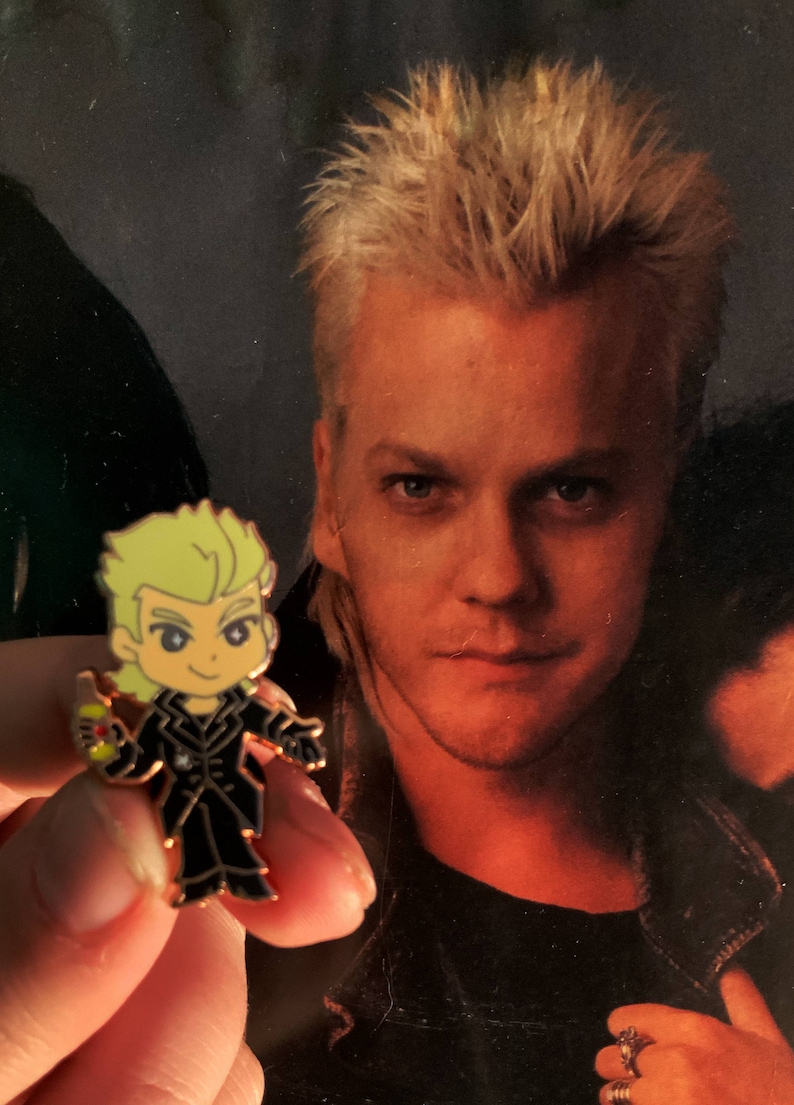 May include: A cartoon-style enamel pin featuring a character with blonde hair and a black outfit. The pin is held in front of a faded image of a man with blonde hair. The pin has a black outline and is filled with yellow, black, and red enamel.