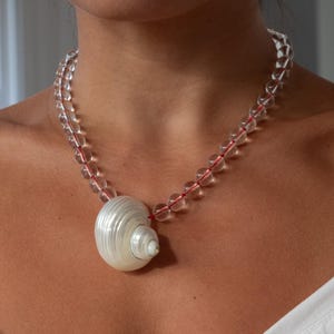 May include: A necklace featuring a single white spiral seashell pendant suspended from a strand of clear, spherical beads. The beads are strung on a thin red cord that is visible through the transparent material of each individual bead.