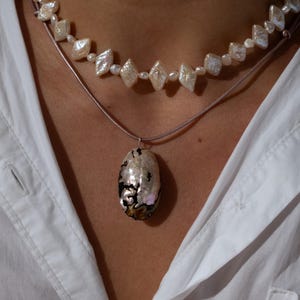 May include: A layered necklace set featuring a choker made of alternating small round and diamond-shaped white iridescent pearls, paired with a second cord necklace holding a large, oval-shaped pendant with a dark, mottled, pearlescent surface.