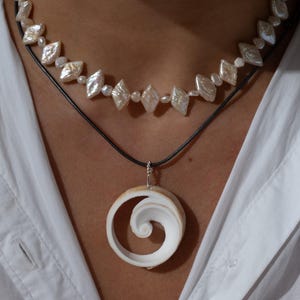 Puede incluir: Un collar de dos hilos con un colgante de concha blanca y un collar de perlas. El colgante tiene un diseño en espiral. El collar superior tiene perlas en forma de diamante. Los collares están en un cordón negro.