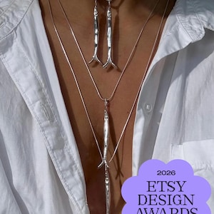 May include: A layered silver necklace set featuring three slender, fish-shaped pendants suspended on delicate chains, worn with a blue and white cord choker. A purple badge in the corner displays the text 2026 ETSY DESIGN AWARDS Finalist.