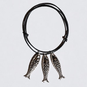 May include: A black cord necklace featuring three silver-toned fish charms hanging from the circular band. The fish pendants have a textured, scale-like surface detail and are suspended from the thin, dark cord which is secured with knots.