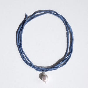 May include: A necklace featuring a single irregular white pearl pendant suspended from a multi-strand blue silk ribbon cord. The cord is wrapped in several loops, creating a layered appearance with a knotted end detail.