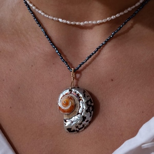 Op de afbeelding: Een ketting met een schelpenhanger. De hanger heeft een spiraalvorm met zilver- en oranjetinten. De bovenste streng is gemaakt van witte parels en de onderste streng van donkere kralen.