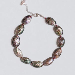 May include: A necklace featuring oval-shaped abalone shell beads with iridescent green, purple, and blue tones. The beads are separated by small red spacer beads and connected by a silver-toned metal chain with a lobster claw clasp.