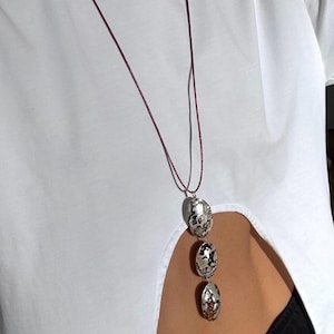 May include: A long necklace featuring a thin, dark maroon cord and three vertically aligned, oval-shaped silver-toned pendants with a textured, hammered surface and dark, irregular mottled patterns across their metallic finish.