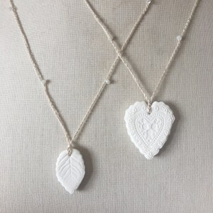 Può includere: Due ciondoli in ceramica bianca, uno a forma di cuore e l'altro a forma di foglia, sono appesi a uno spago bianco con perline bianche. I ciondoli hanno disegni intricati.