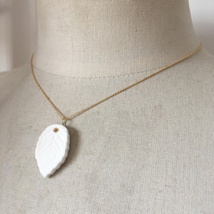 Puede incluir: Un delicado collar de cadena dorada con un colgante de hoja de porcelana blanca. La hoja tiene un patrón de nervaduras detallado y un pequeño acento dorado.