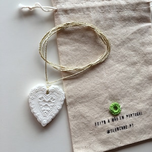 May include: White ceramic heart pendant with intricate detailing, hanging from a gold cord. The pendant is packaged in a natural canvas bag with the text "FEITO A MÃO EM PORTUGAL @SLOWCARE.PT" printed on it.