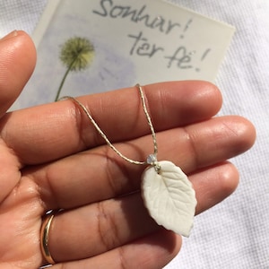 Puede incluir: Un delicado collar con un colgante de hoja blanca y una cadena dorada. El colgante de hoja tiene vetas detalladas. El collar se sostiene en una mano, con un anillo dorado en el dedo. Una tarjeta con el texto "Sonhar! ter fé!" está en el fondo.