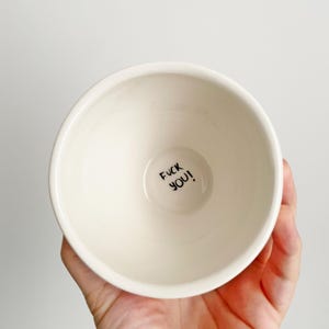 Puede incluir: Un cuenco de cer&aacute;mica color crema con el texto "FUCK YOU!" en letras negras en la base. El cuenco se sostiene en una mano sobre un fondo blanco. El cuenco tiene un dise&ntilde;o simple y minimalista.