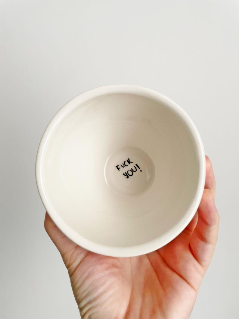 Puede incluir: Un cuenco de cer&aacute;mica color crema con el texto "FUCK YOU!" en letras negras en la base. El cuenco se sostiene en una mano sobre un fondo blanco. El cuenco tiene un dise&ntilde;o simple y minimalista.