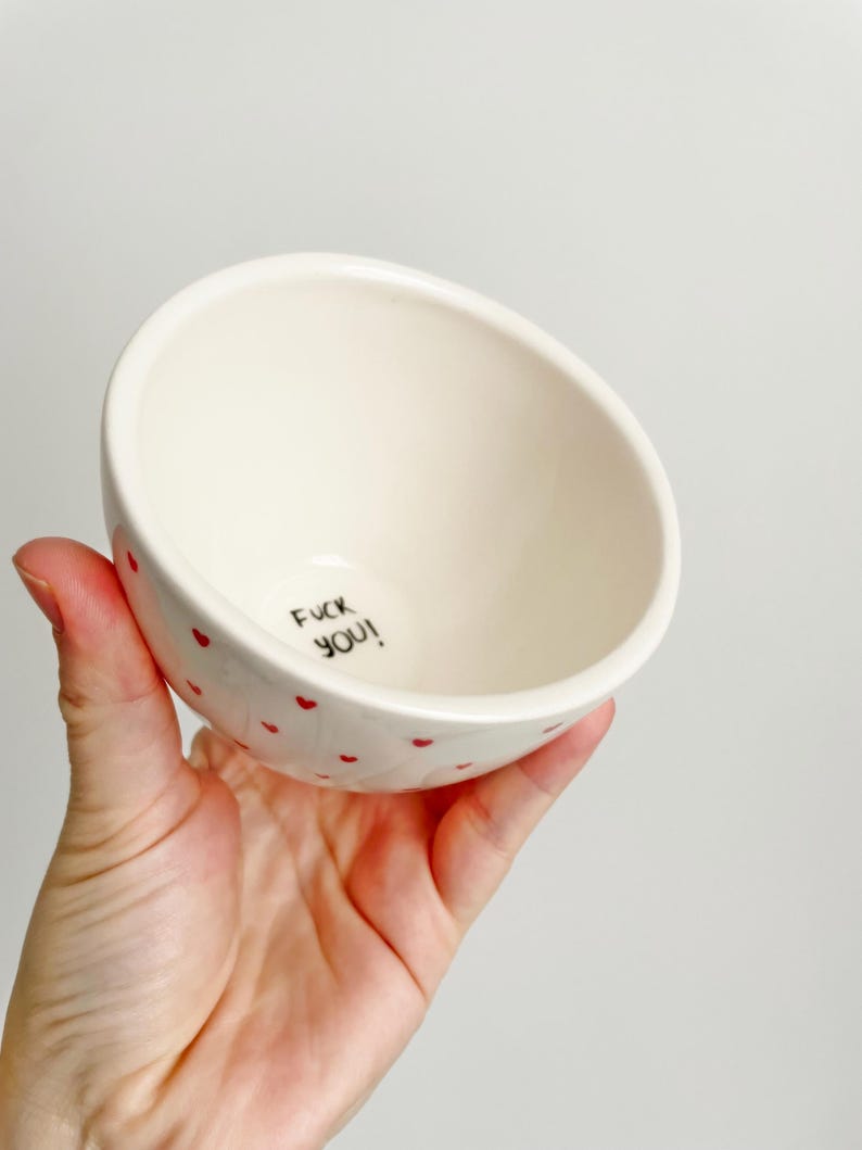 Puede incluir: Un cuenco de cer&aacute;mica blanco con dise&ntilde;os en forma de coraz&oacute;n rojo. El cuenco tiene el texto "FUCK YOU!" escrito en negro en su interior. El cuenco est&aacute; sostenido en una mano.