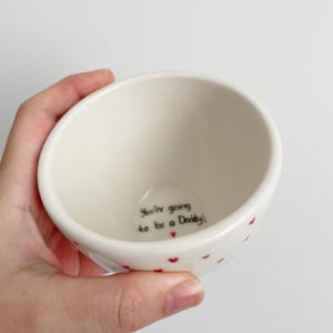 Puede incluir: Un peque&ntilde;o cuenco de cer&aacute;mica blanquecina con detalles de corazones rojos. El cuenco tiene el texto "You're going to be a Daddy!" escrito en escritura negra en el interior. El cuenco est&aacute; sostenido en una mano.