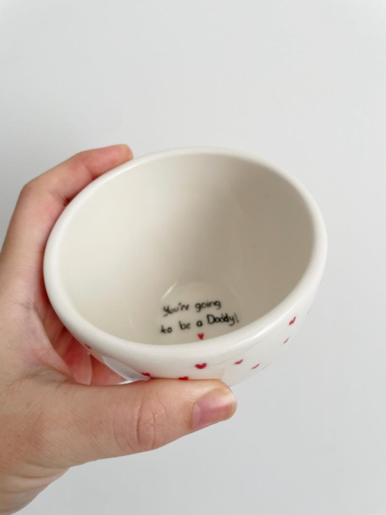 Puede incluir: Un peque&ntilde;o cuenco de cer&aacute;mica blanquecina con detalles de corazones rojos. El cuenco tiene el texto "You're going to be a Daddy!" escrito en escritura negra en el interior. El cuenco est&aacute; sostenido en una mano.
