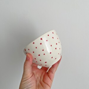 Puede incluir: Un cuenco de cer&aacute;mica blanca con acabado brillante, adornado con peque&ntilde;os dise&ntilde;os de corazones rojos espaciados. El cuenco se sostiene en &aacute;ngulo, mostrando su interior y el patr&oacute;n. El fondo es gris claro liso.