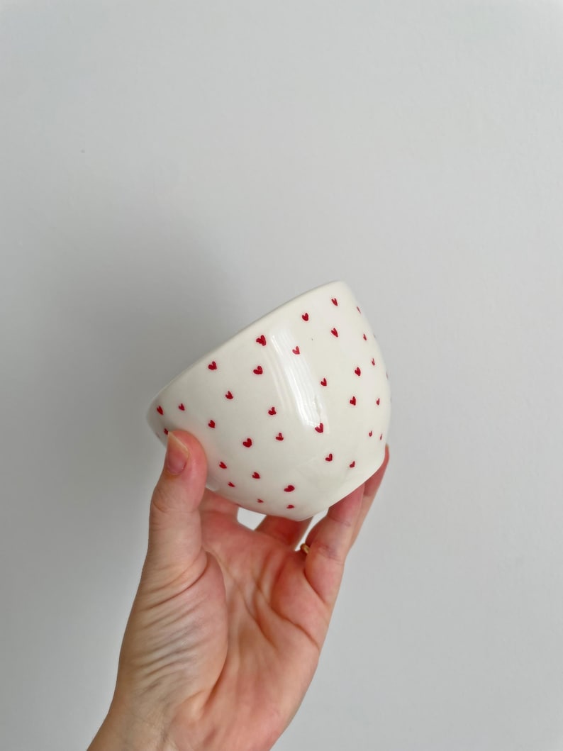 Puede incluir: Un cuenco de cer&aacute;mica blanca con acabado brillante, adornado con peque&ntilde;os dise&ntilde;os de corazones rojos espaciados. El cuenco se sostiene en &aacute;ngulo, mostrando su interior y el patr&oacute;n. El fondo es gris claro liso.