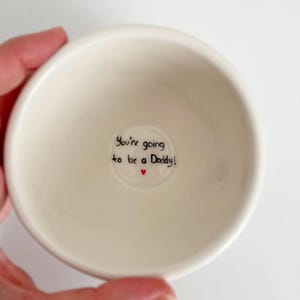 Puede incluir: Un peque&ntilde;o cuenco de cer&aacute;mica blanquecina con el texto "You're going to be a Daddy!" en escritura negra y un peque&ntilde;o coraz&oacute;n rojo. El cuenco est&aacute; sostenido por una mano, probablemente un regalo.