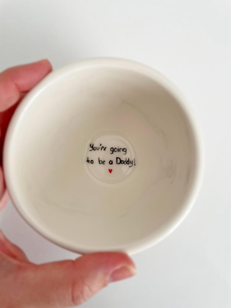 Puede incluir: Un peque&ntilde;o cuenco de cer&aacute;mica blanquecina con el texto "You're going to be a Daddy!" en escritura negra y un peque&ntilde;o coraz&oacute;n rojo. El cuenco est&aacute; sostenido por una mano, probablemente un regalo.