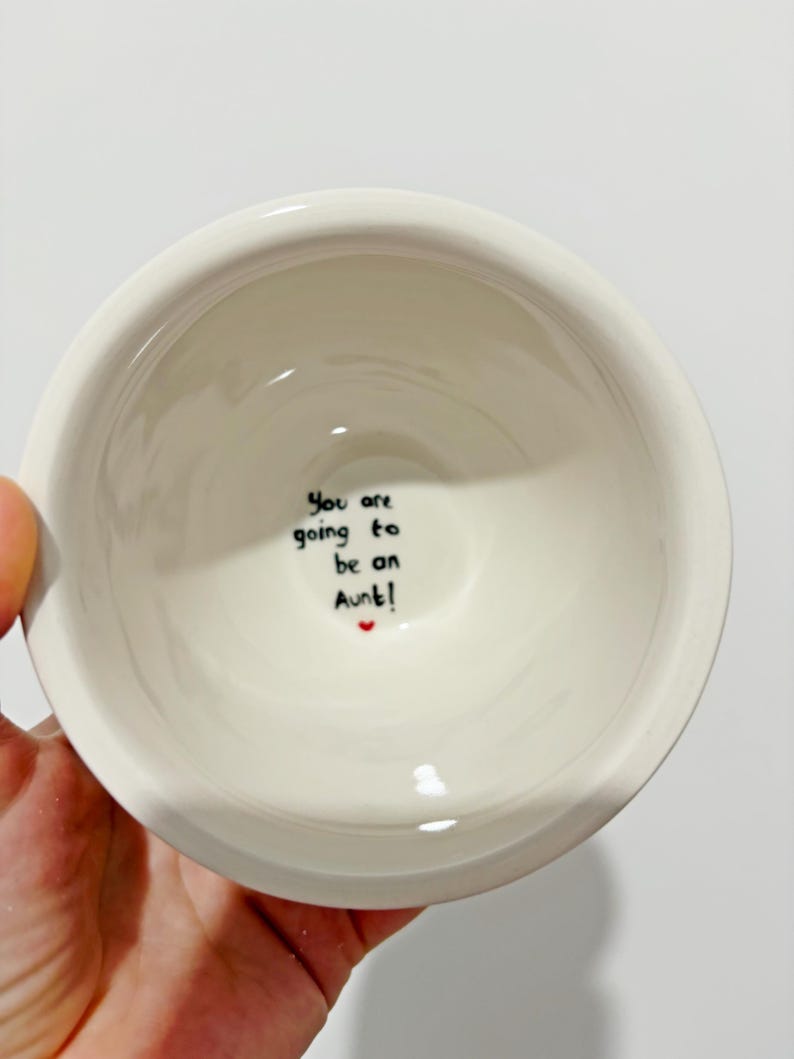 Puede incluir: Un cuenco de cer&aacute;mica blanco con el texto escrito a mano "You are going to be an Aunt!" en tinta negra, con un peque&ntilde;o coraz&oacute;n rojo. El cuenco se sostiene en una mano sobre un fondo blanco.