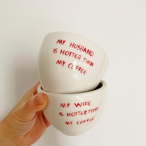 Może przedstawiać: Dwa białe ceramiczne kubki do kawy ułożone jeden na drugim. Na górnym kubku znajduje się czerwony napis "MY HUSBAND IS HOTTER THAN MY COFFEE". Na dolnym kubku znajduje się czerwony napis "MY WIFE IS HOTTER THAN MY COFFEE". Kubki są trzymane przez dłoń.