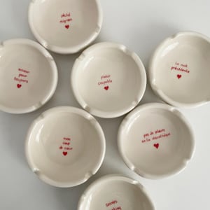 May include: Off-white ceramic ashtrays with red text and heart illustrations. Each ashtray has a different French phrase, such as "péché mignon" and "amour pour toujours". The ashtrays are round with a small lip.