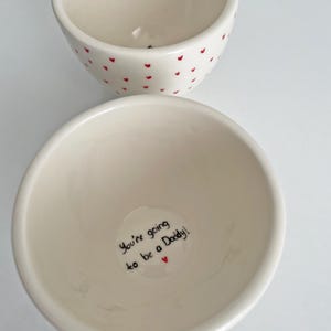 Puede incluir: Dos cuencos de cer&aacute;mica blanquecina. Uno est&aacute; decorado con peque&ntilde;os corazones rojos. El otro tiene el texto "You're going to be a Daddy!" en escritura negra con un peque&ntilde;o coraz&oacute;n rojo. Los cuencos son probablemente para servir comida o como objetos decorativos.