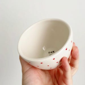 Puede incluir: Un peque&ntilde;o cuenco de cer&aacute;mica blanquecina con detalles de corazones rojos. La palabra "FUCK" est&aacute; impresa en negro en el interior. El cuenco est&aacute; sostenido en una mano.