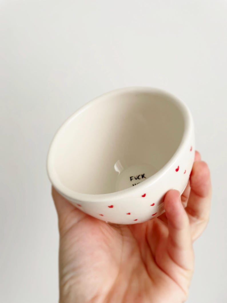 Puede incluir: Un peque&ntilde;o cuenco de cer&aacute;mica blanquecina con detalles de corazones rojos. La palabra "FUCK" est&aacute; impresa en negro en el interior. El cuenco est&aacute; sostenido en una mano.