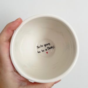 Puede incluir: Un cuenco de cer&aacute;mica blanco con el texto "You're going to be a Daddy!" en escritura negra y un peque&ntilde;o coraz&oacute;n rojo. El cuenco tiene un dise&ntilde;o simple y redondeado.