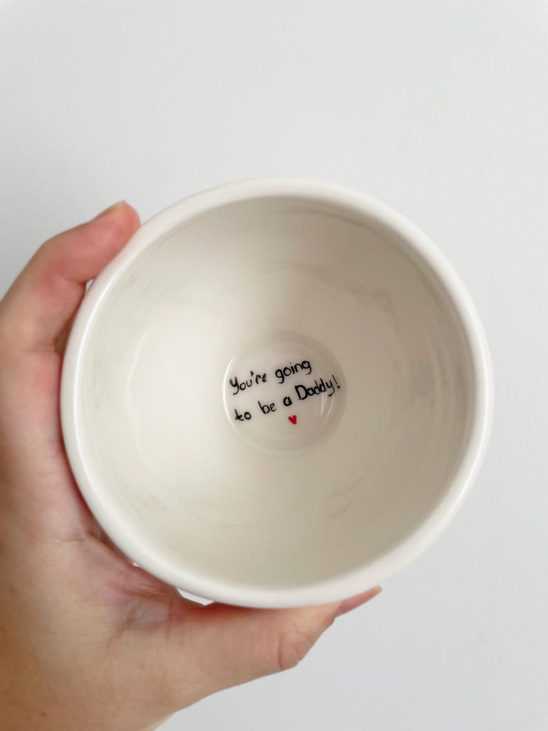 Puede incluir: Un cuenco de cer&aacute;mica blanco con el texto "You're going to be a Daddy!" en escritura negra y un peque&ntilde;o coraz&oacute;n rojo. El cuenco tiene un dise&ntilde;o simple y redondeado.