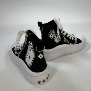 May include: Black high-top sneakers with white soles and laces. The sneakers feature playing card and dice graphics, along with decorative chains and charms. The heels are adorned with heart, spade, and diamond accents.