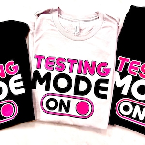 Op de afbeelding: Drie T-shirts met de tekst "Testing Mode On" in witte letters met een roze omtrek. De tekst staat op een zwart, grijs en zwart T-shirt. De "On" is een roze cirkel met een witte omtrek.