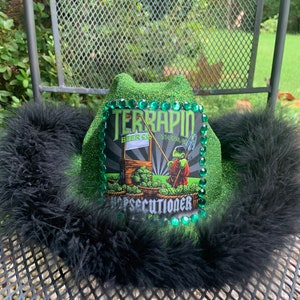 May include: Green glitter cowboy hat with a black feather trim. The hat features a Terrapin Beer Co. logo with a green hops graphic and the text "Hopsecutioner".