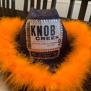 May include: A black sparkly cowboy hat with a Knob Creek Kentucky Straight Bourbon Whiskey label with rhinestones around the label. The hat is decorated with a bright orange feather boa.