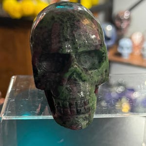 May include: A carved skull figurine made of green and maroon stone. The skull has detailed features, including eye sockets, a nose cavity, and teeth. The stone has a polished, reflective surface. The skull is displayed on a clear glass shelf.