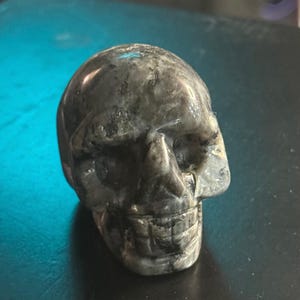 May include: A carved stone skull in shades of gray and black. The skull has detailed features, including eye sockets, a nose, and teeth. The stone has a polished, smooth surface.