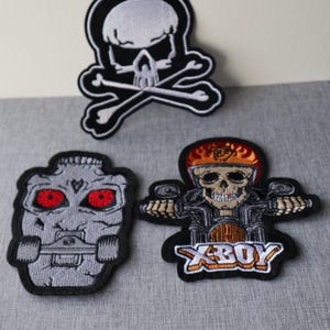 Peut inclure: Trois écussons brodés avec des motifs de crânes. L'un représente un crâne et des os croisés, un autre un crâne sur un skateboard, et le troisième un crâne à moto avec le texte "XBOY". Tous les écussons ont des bordures noires.