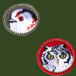 May include: Two embroidered patches with bird designs. One shows a white bird with red details on a grey background. The other depicts an owl with yellow eyes on a red background, both with gold trim.