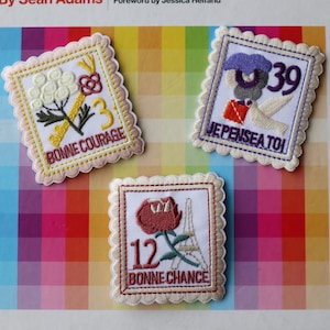 May include: Three embroidered patches with scalloped edges. The first patch is pink and white with a flower and key design and the text "Bonne Courage" and the number "3". The second patch is blue and white with a bird design and the text "Je Pense a Toi" and the number "39". The third patch is white and red with a flower and Eiffel Tower design and the text "Bonne Chance" and the number "12".