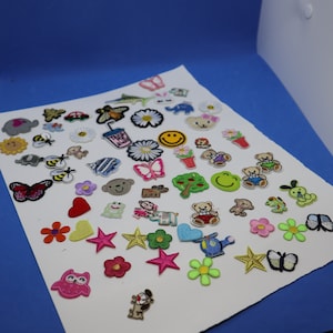 May include: A selection of colourful embroidered patches with diverse designs. Includes flowers, stars, butterflies, smiley faces, animals, and a coffee cup. Patches are in various colours, including red, yellow, pink, green, and blue, arranged on a white surface.