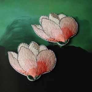 May include: Two embroidered pink and white flower patches with green centers. The flowers are on a green background.