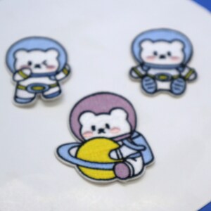 Peut inclure: Trois patchs brodés thermocollants représentant un ours polaire en dessin animé portant une combinaison d'astronaute. L'ours au centre tient une planète jaune avec un anneau bleu autour. Les deux autres ours portent des combinaisons d'astronaute bleues avec des accents jaunes.