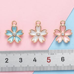 May include: Five gold-tone metal charms shaped like cherry blossoms. Each charm has a different colored enamel finish: black, light blue, white, pink, and pink with a pearl center. The charms are approximately 1.5 cm in diameter and are shown next to a ruler.