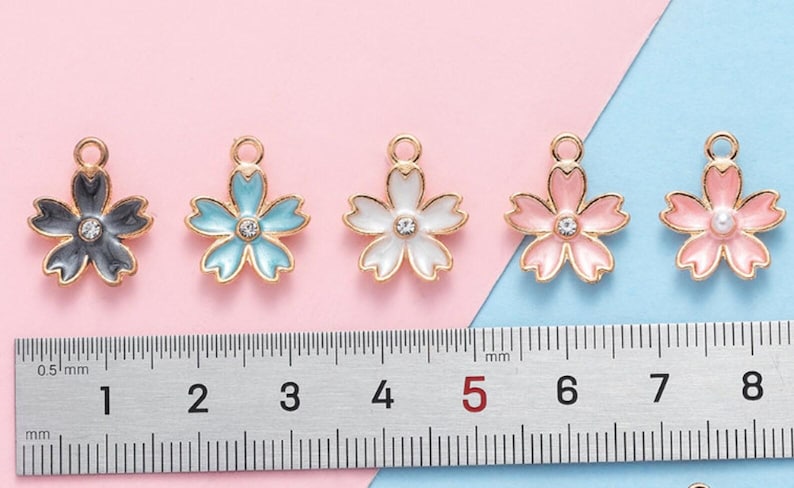 May include: Five gold-tone metal charms shaped like cherry blossoms. Each charm has a different colored enamel finish: black, light blue, white, pink, and pink with a pearl center. The charms are approximately 1.5 cm in diameter and are shown next to a ruler.