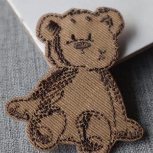 May include: A tan fabric patch shaped like a seated teddy bear, featuring dark brown embroidered outlines and details that define the bear's ears, face, limbs, and torso, with a textured twill weave visible throughout the material.