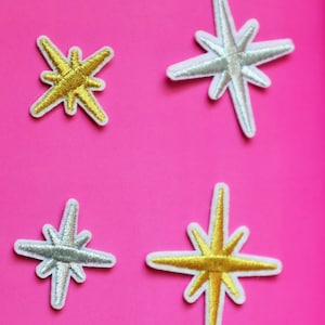 May include: Four iron-on patches in the shape of stars. Two are gold and two are silver. The stars have a white outline.