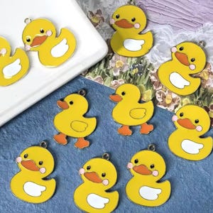 Peut inclure: Plusieurs breloques en forme de canard jaune avec des becs et des pieds orange et des ventres blancs. Chaque breloque a une petite boucle en haut pour l'attacher à des bijoux ou à d'autres créations. Les breloques sont sur une surface bleue.